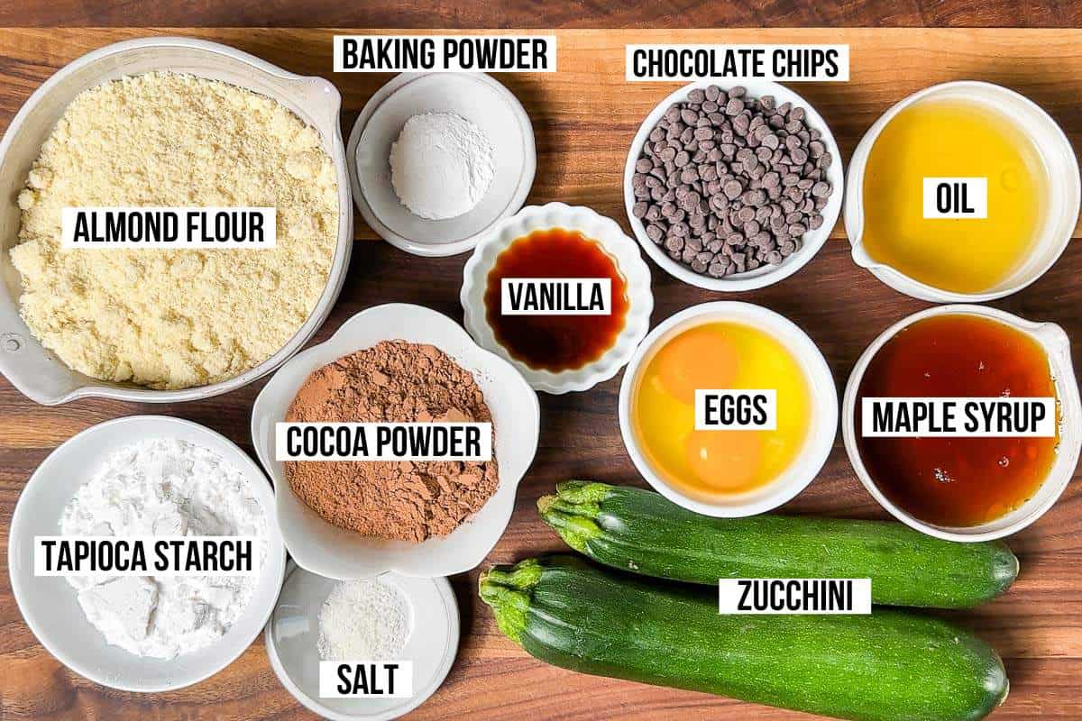 All the ingredients for Healthy Chocolate Zucchini Muffins: almond flour, tapioca starch, cocoa powder, zucchini, vanilla, baking powder, chocolate chips, maple syrup, eggs, avocado oil, and salt in bowls on a wood cutting board.