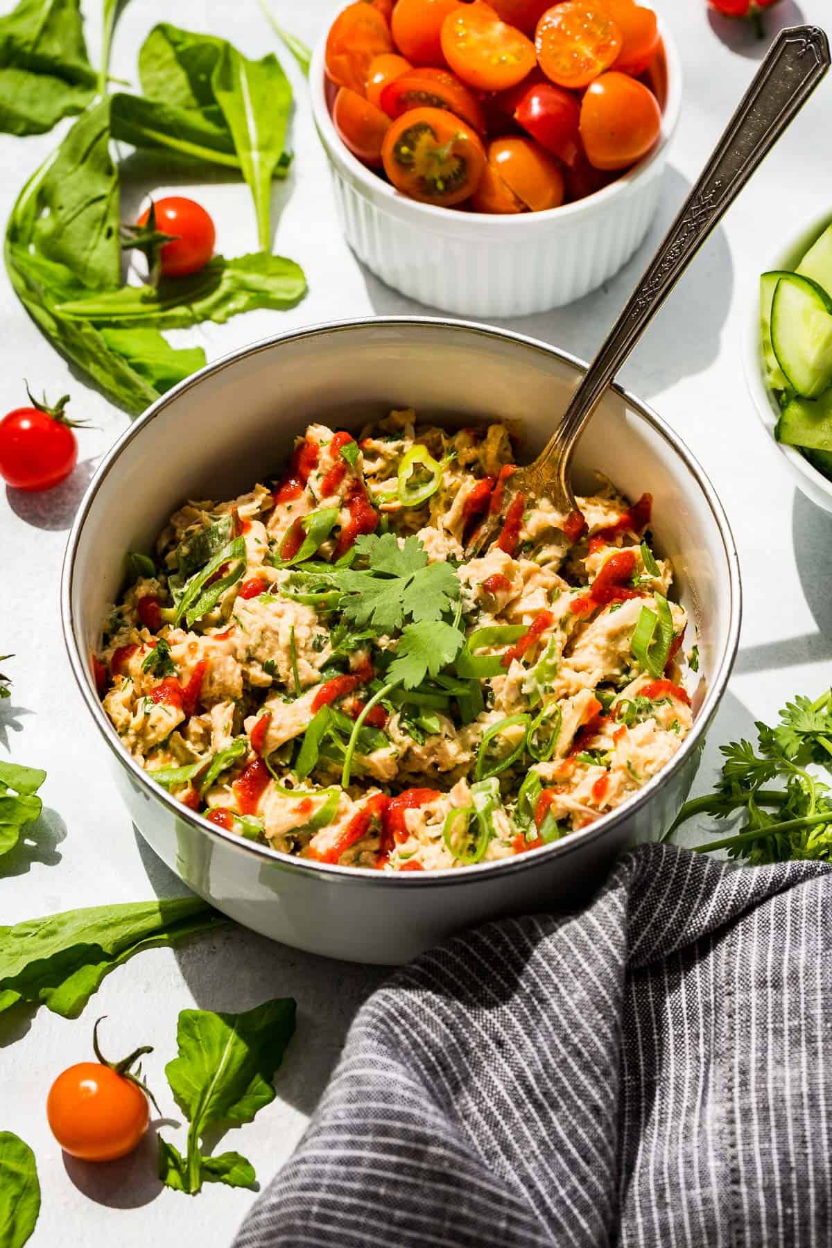 A white bowl with Spicy Tuna Salad topped with sriracha and cilantro with greens and cherry tomatoes around it.