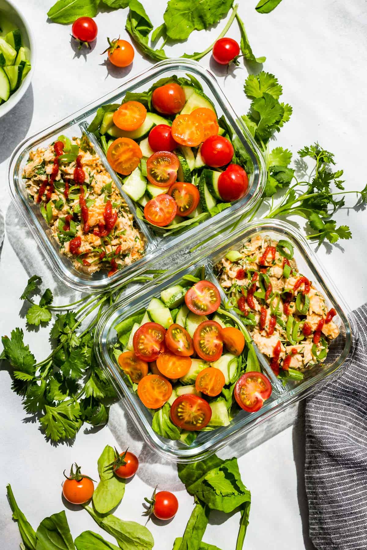 Spicy Tuna Salad in meal prep containers place on a blue background on a diagonal with cherry tomatoes and cilantro around it.