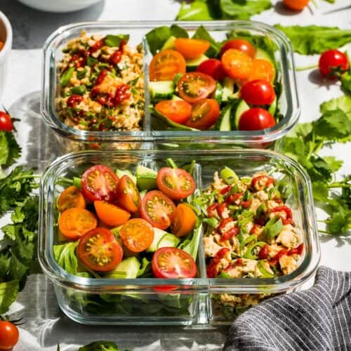 Side view of Spicy Tuna Salad in meal prep containers with cherry tomatoes and cucumbers on the side with greens and cilantro around the containers.