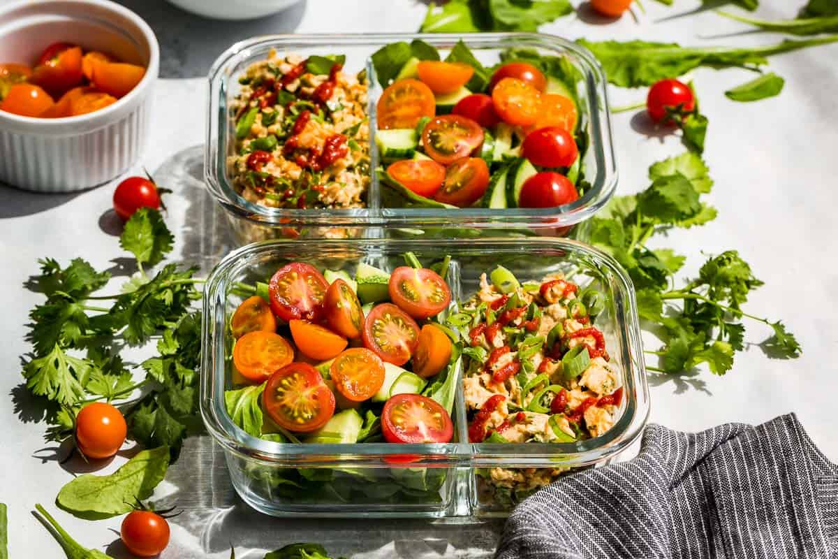 Side view of Spicy Tuna Salad in meal prep containers with cherry tomatoes and cucumbers on the side with greens and cilantro around the containers.