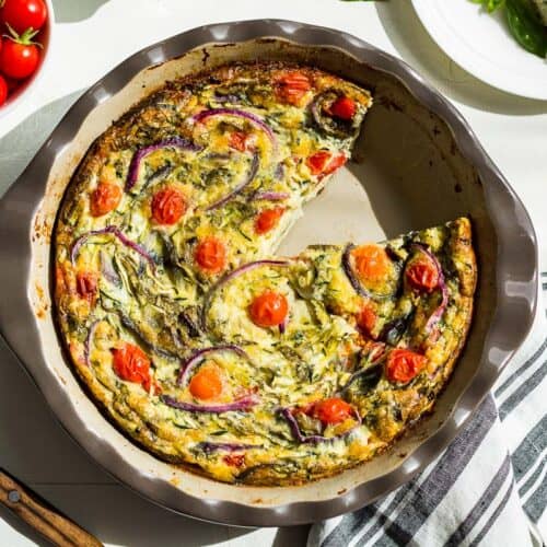 Straight down view of vegetarian breakfast casserole in a pie plate with a slice take out and cherry tomatoes scattered around.