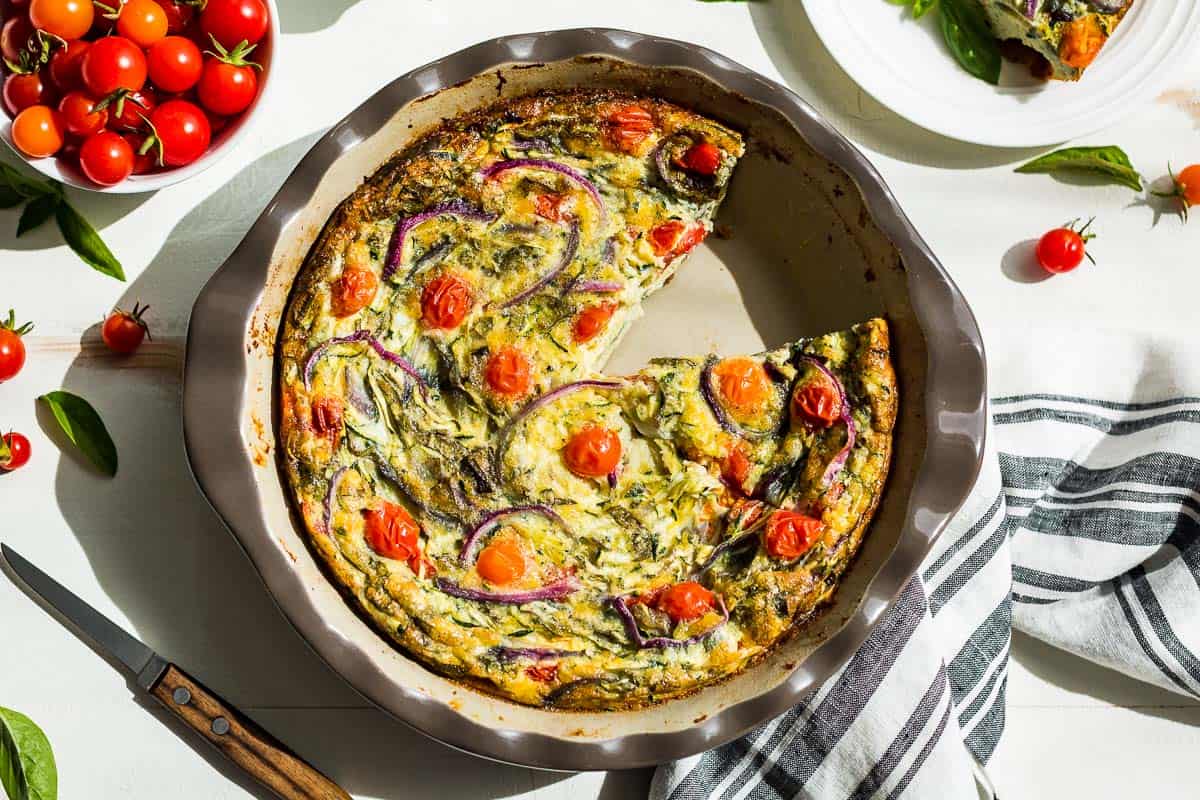 Straight down view of vegetarian breakfast casserole in a pie plate with a slice take out and cherry tomatoes scattered around.