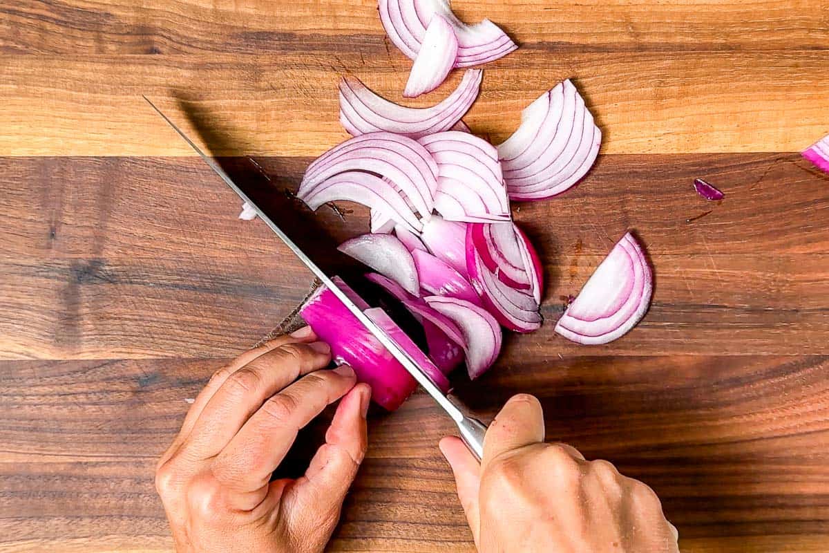 Cutting the red onion into half moon slices with a chefs knife on a wood cutting board.