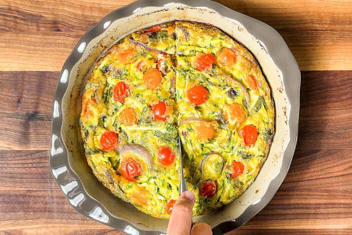 Cutting the baked vegetarian breakfast casserole with a knife on a wood cutting board.