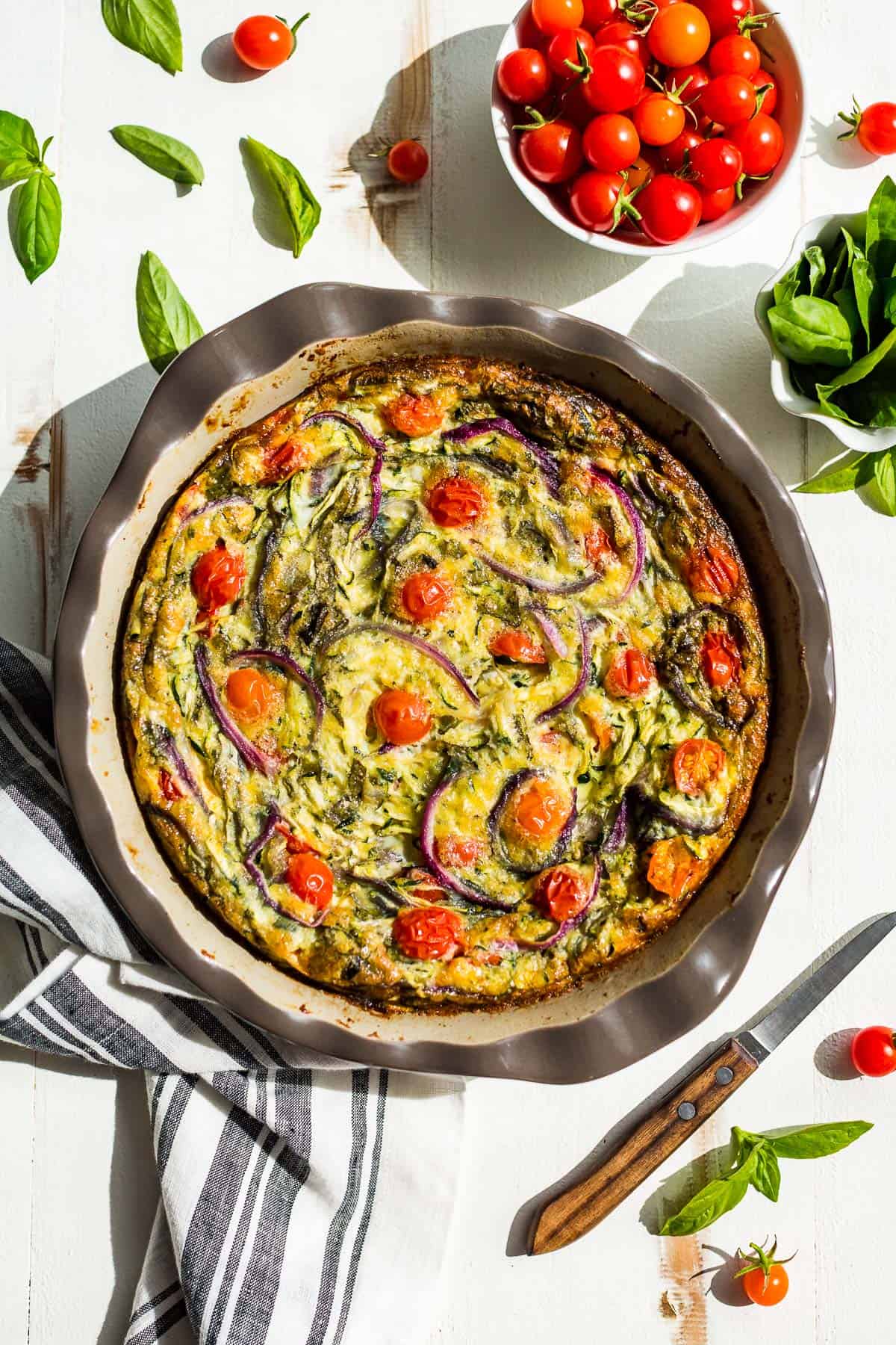 Straight down view of vegetarian breakfast casserole in a pie plate with cherry tomatoes and basil in the background.