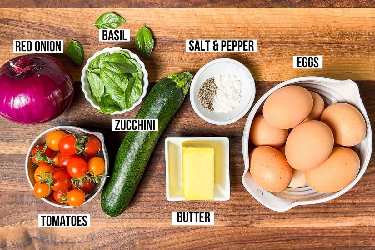 Ingredients for the Vegetarian breakfast casserole, eggs, butter, zucchini, red onion, basil, tomatoes, salt and pepper in bowls on a wood cutting board.