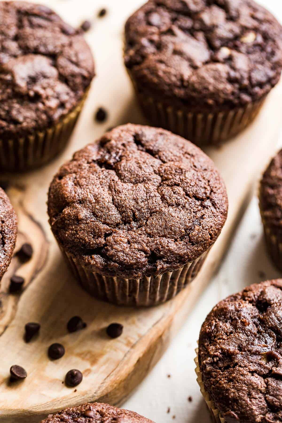Side view of healthy Chocolate Zucchini Muffins on a wood board with chocolate chips scattered around.