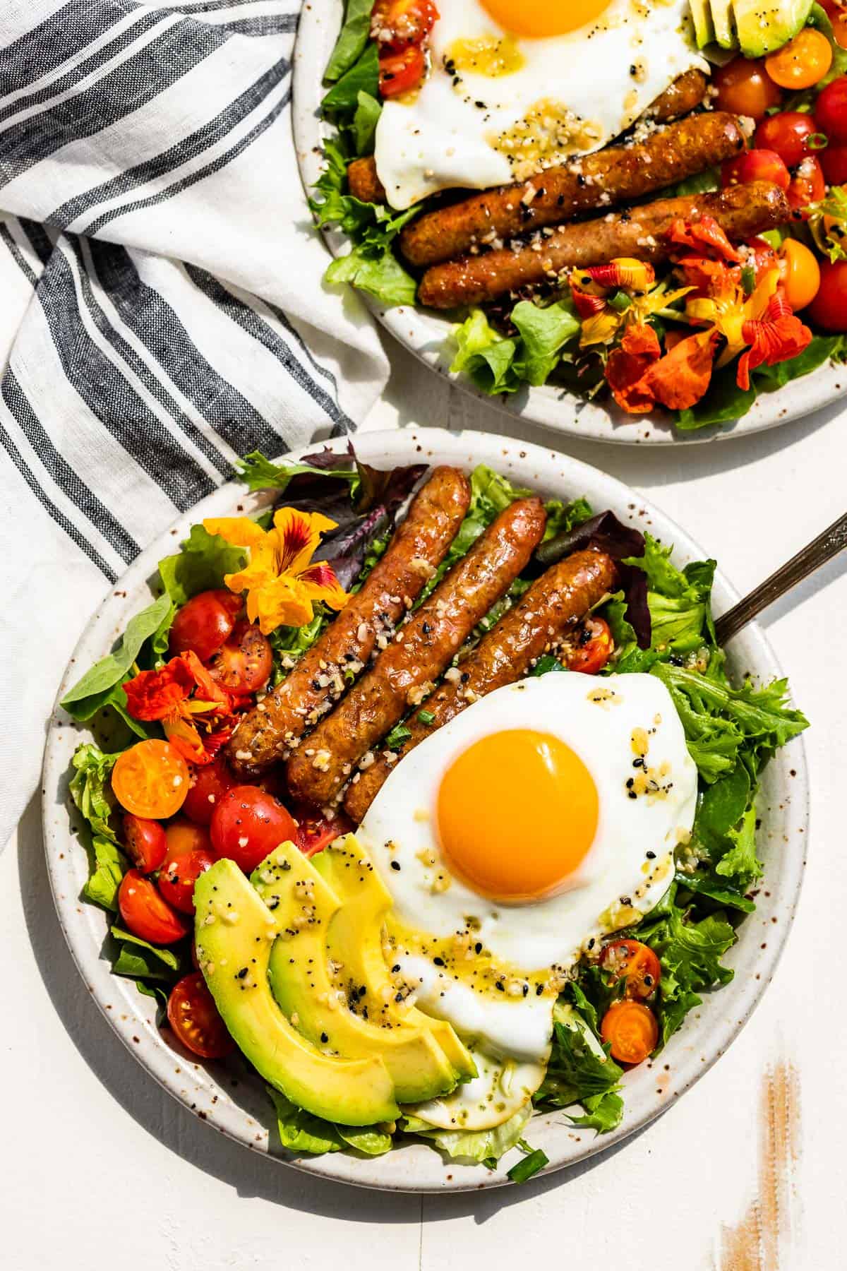 Straight down view of two breakfast salads on pottery plates on a white background with a blue and white striped linen in the background.