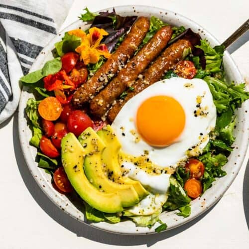 Straight down view of a pottery plate filled with breakfast salad topped with an over easy egg, sliced avocado, cherry tomatoes, and sausages on a white background.