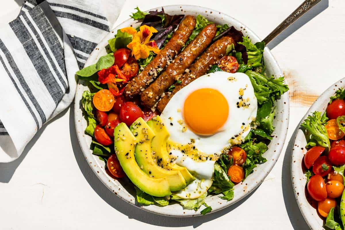 Straight down view of a pottery plate filled with breakfast salad topped with an over easy egg, sliced avocado, cherry tomatoes, and sausages on a white background.