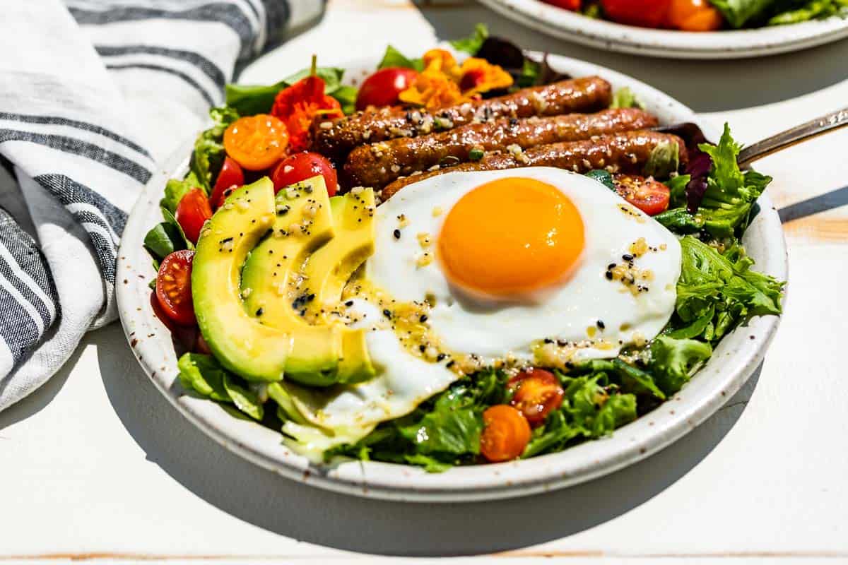 Side view of breakfast salad topped with an over easy egg, sliced avocado, and sausages on a white background.