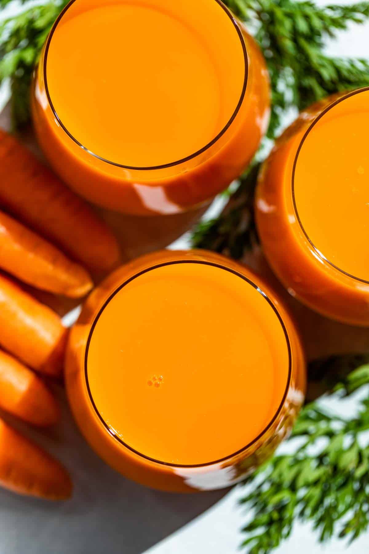 Straight down view of carrot juice in three glasses with carrots and carrot tops on the side.