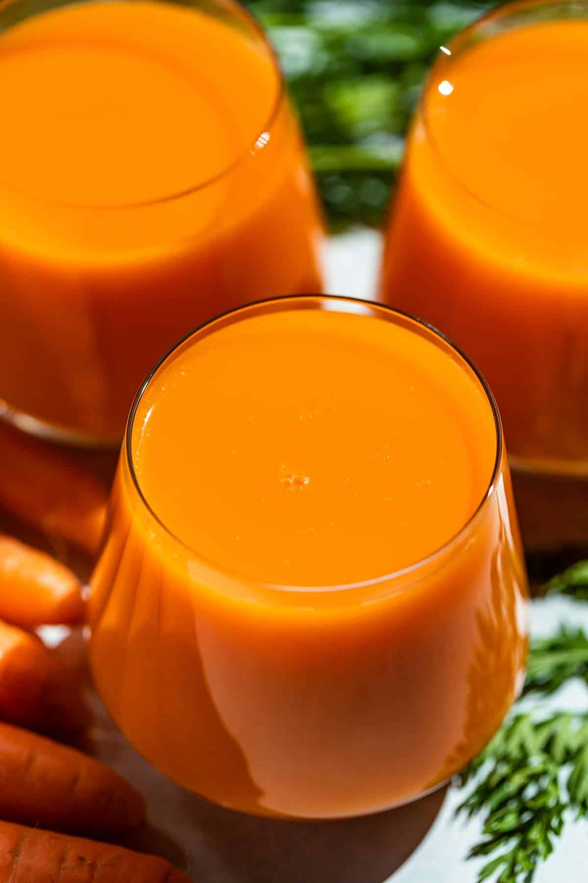 Partially downwards view of carrot juice in three glasses with carrot tops in the background.