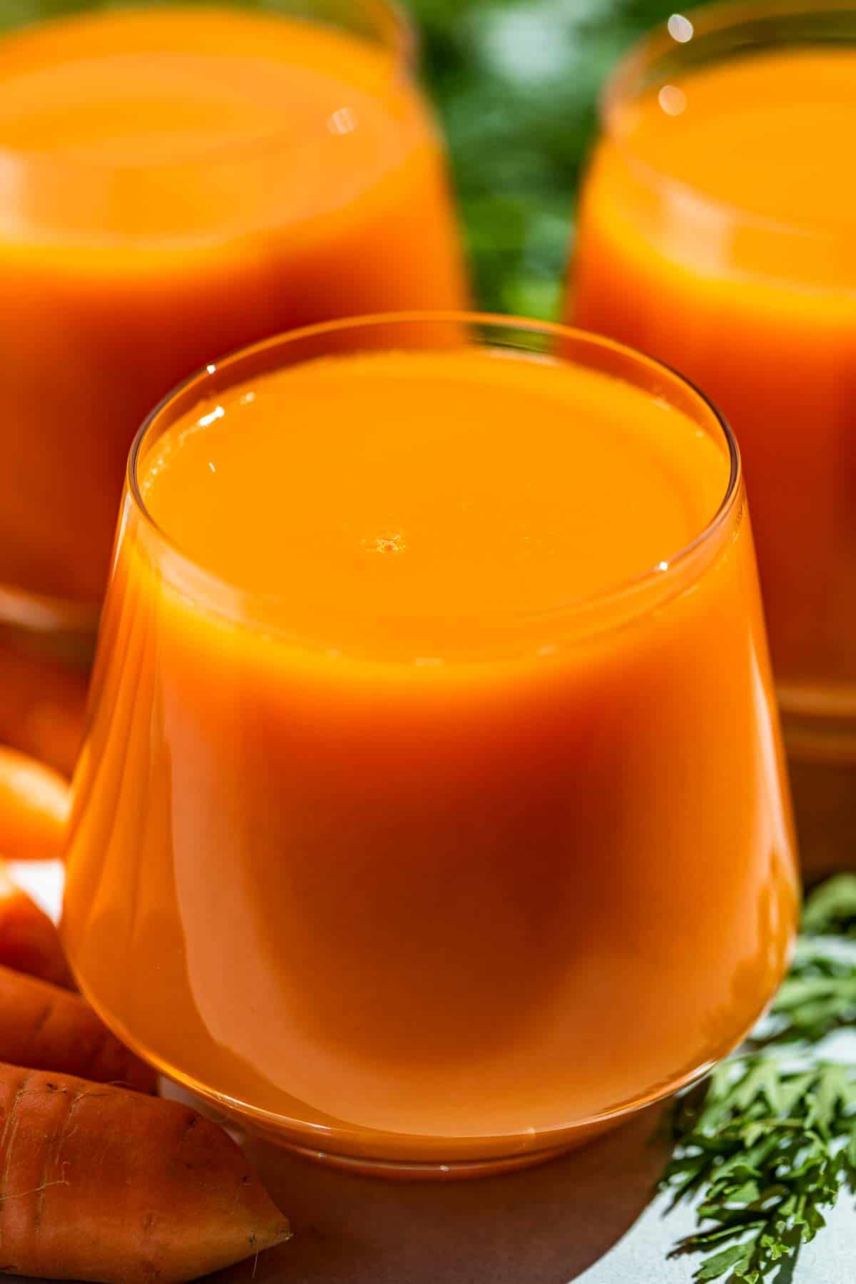 Side view of carrot juice in three glasses with carrots and carrot tops on the side.