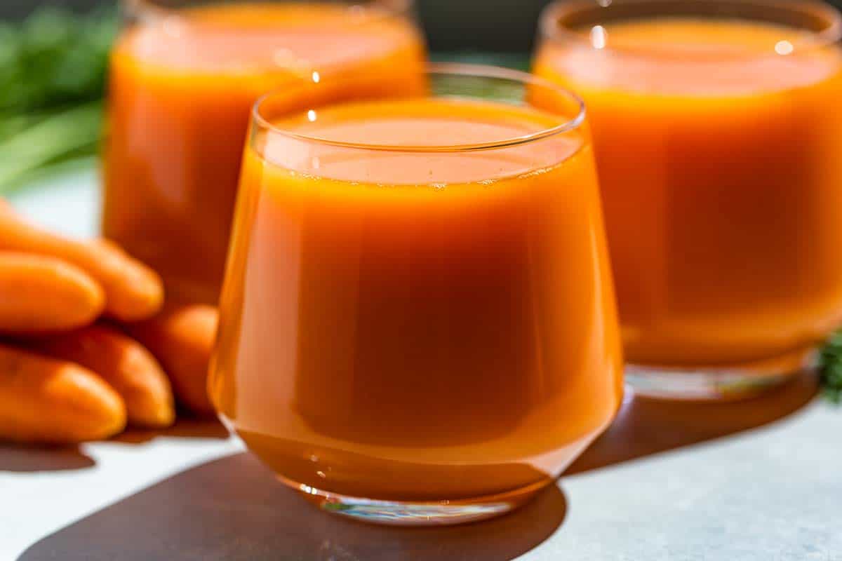 Side view of carrot juice in three glasses with carrots on the side on a blue background.