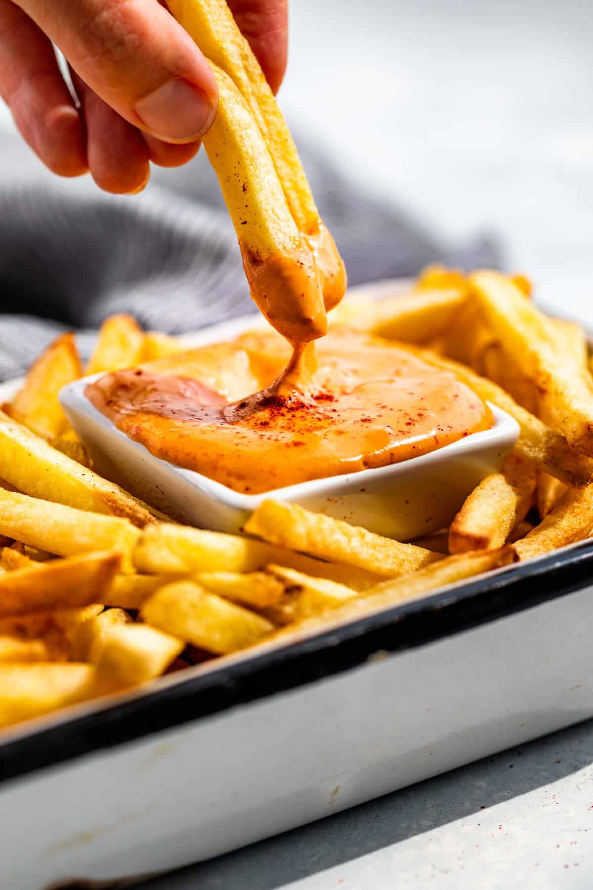 A hand dipping two French fries into a square dish of Chipotle Aioli.