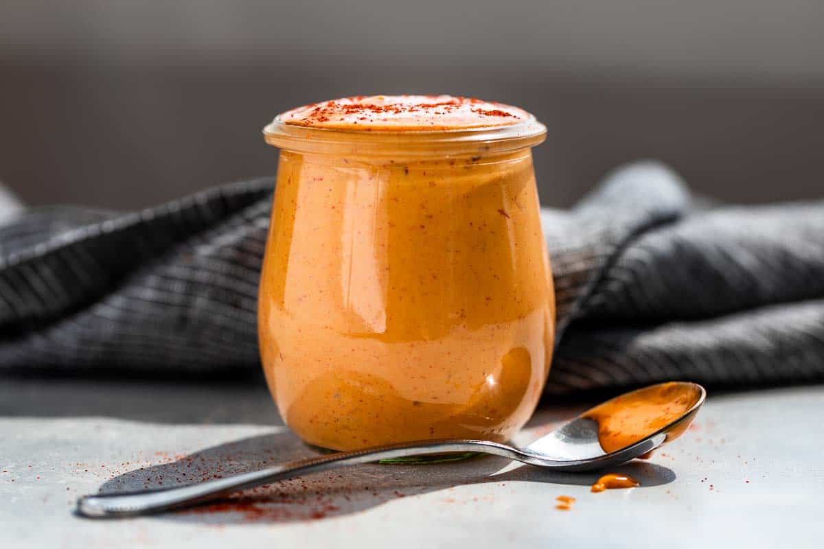 Side view of Chipotle Aioli in a glass jar on a blue background with a silver spoon sitting in front of the container.