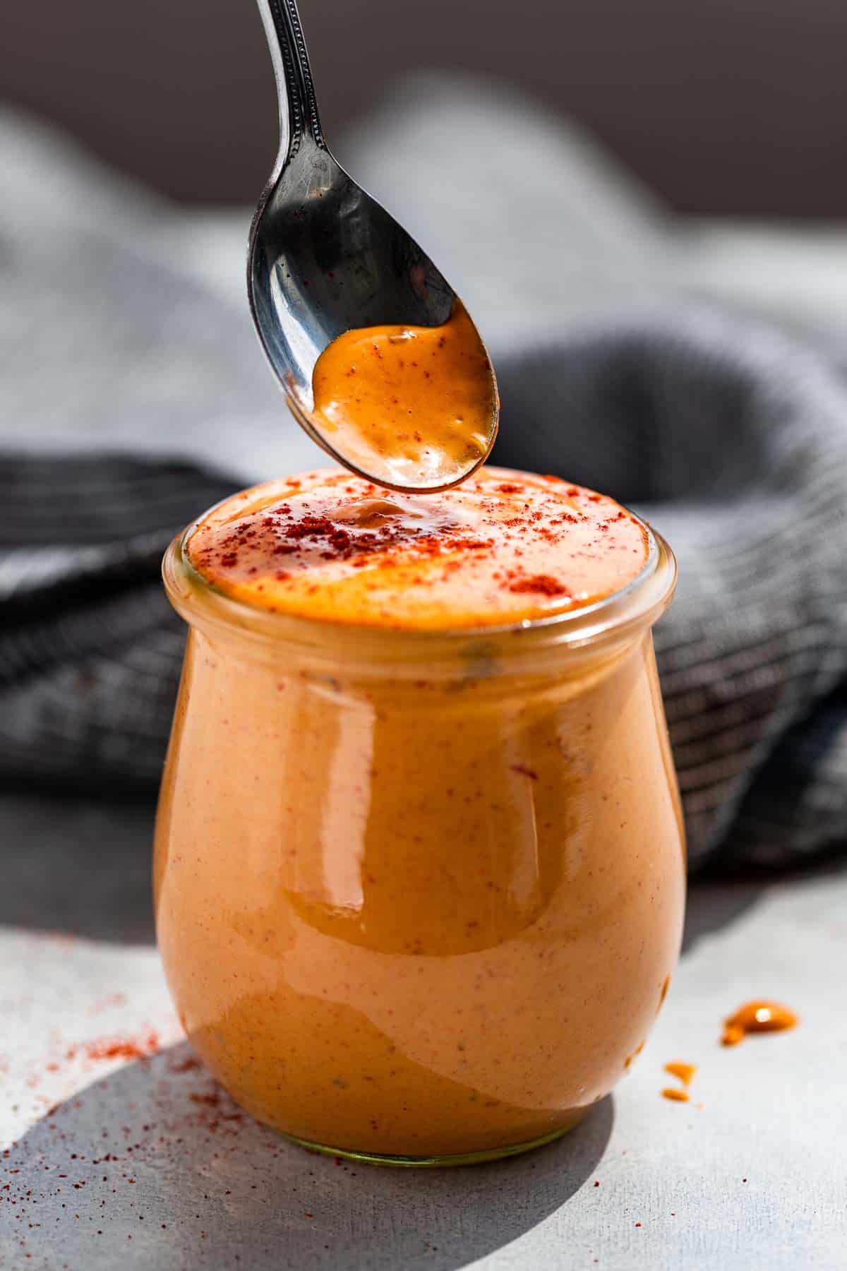 A glass jar filled with Chipotle Aioli with a silver spoon scooping some out on a blue background.