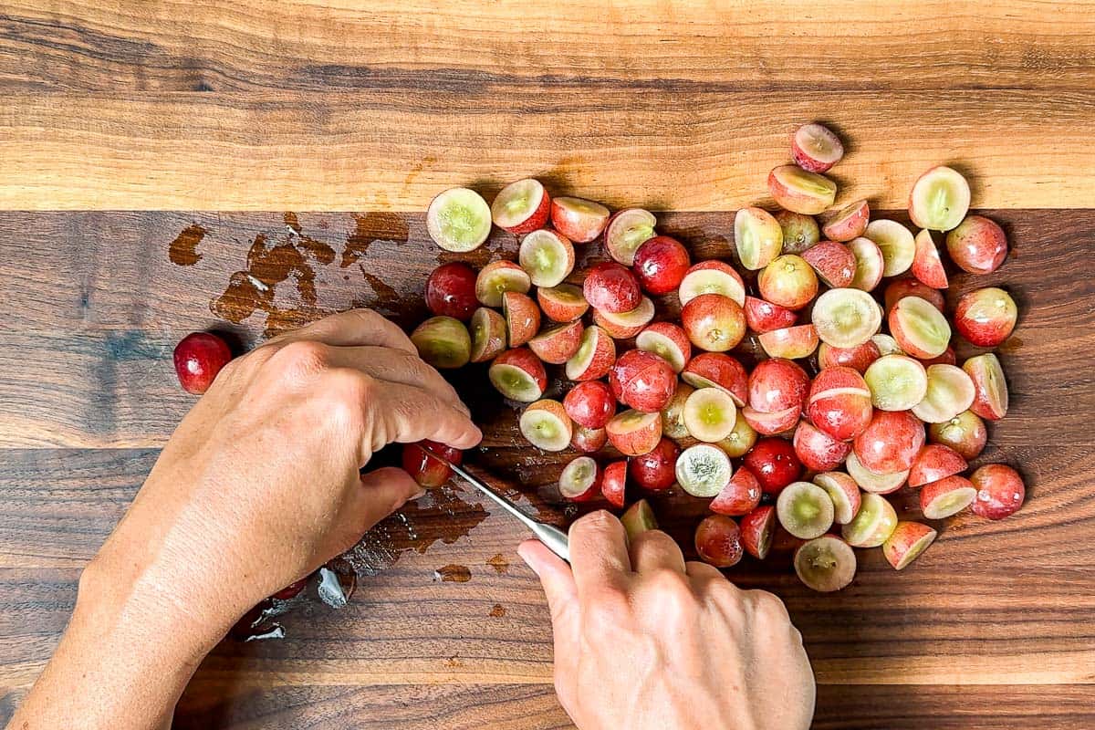 Cutting the red grapes in half with a pairing knife on a wood cutting board.