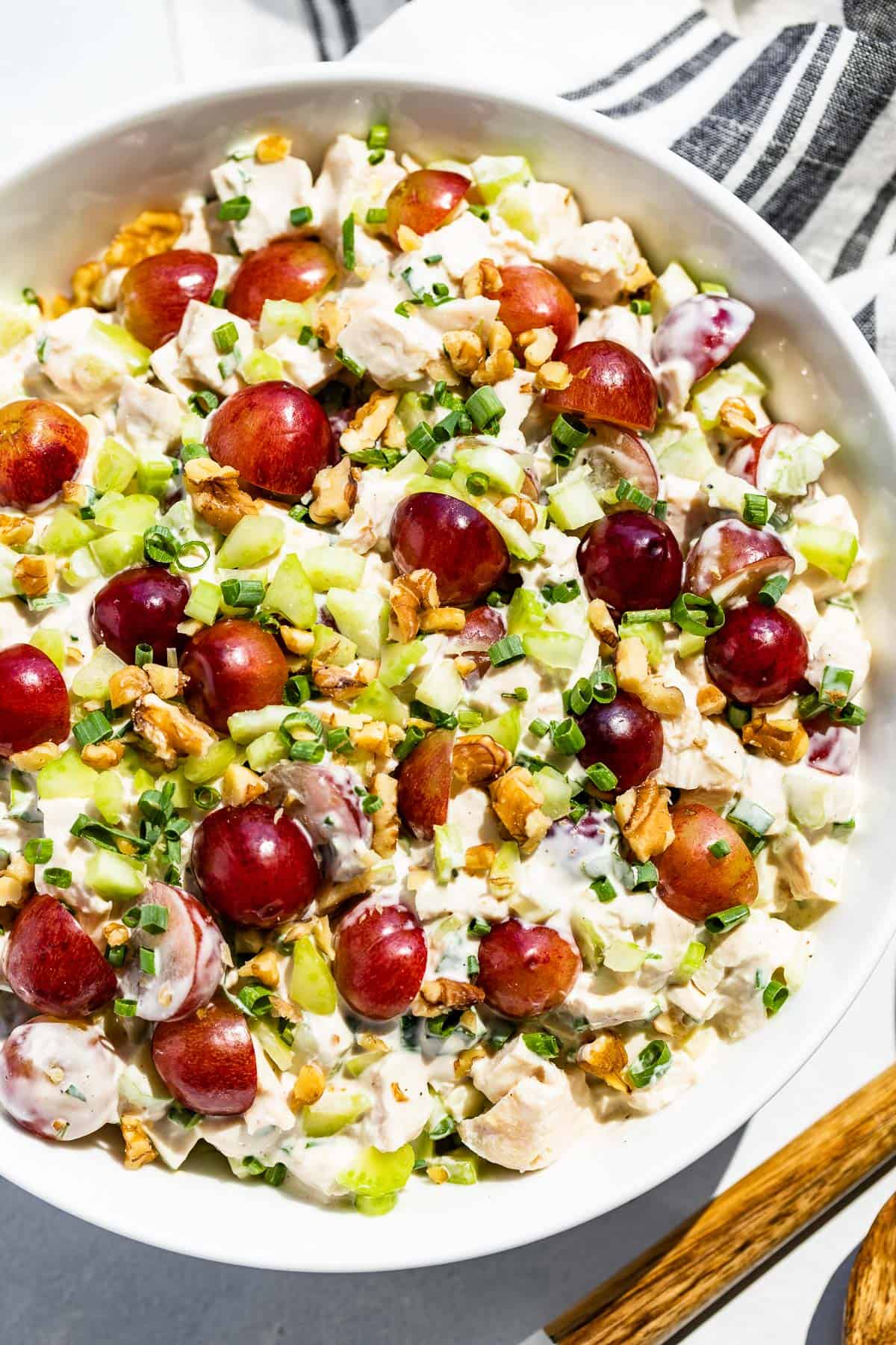 Close up straight down view of healthy chicken salad in a white bowl topped with grapes and wood serving spoons on the side.