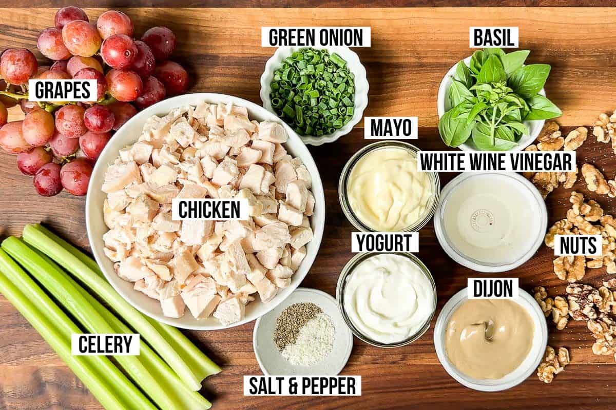 Grapes, celery, cooked chicken, green onion, Greek yogurt, mayonnaise, Dijon mustard, basil, walnuts, white wine vinegar, sea salt and pepper in bowls on a wood cutting board.