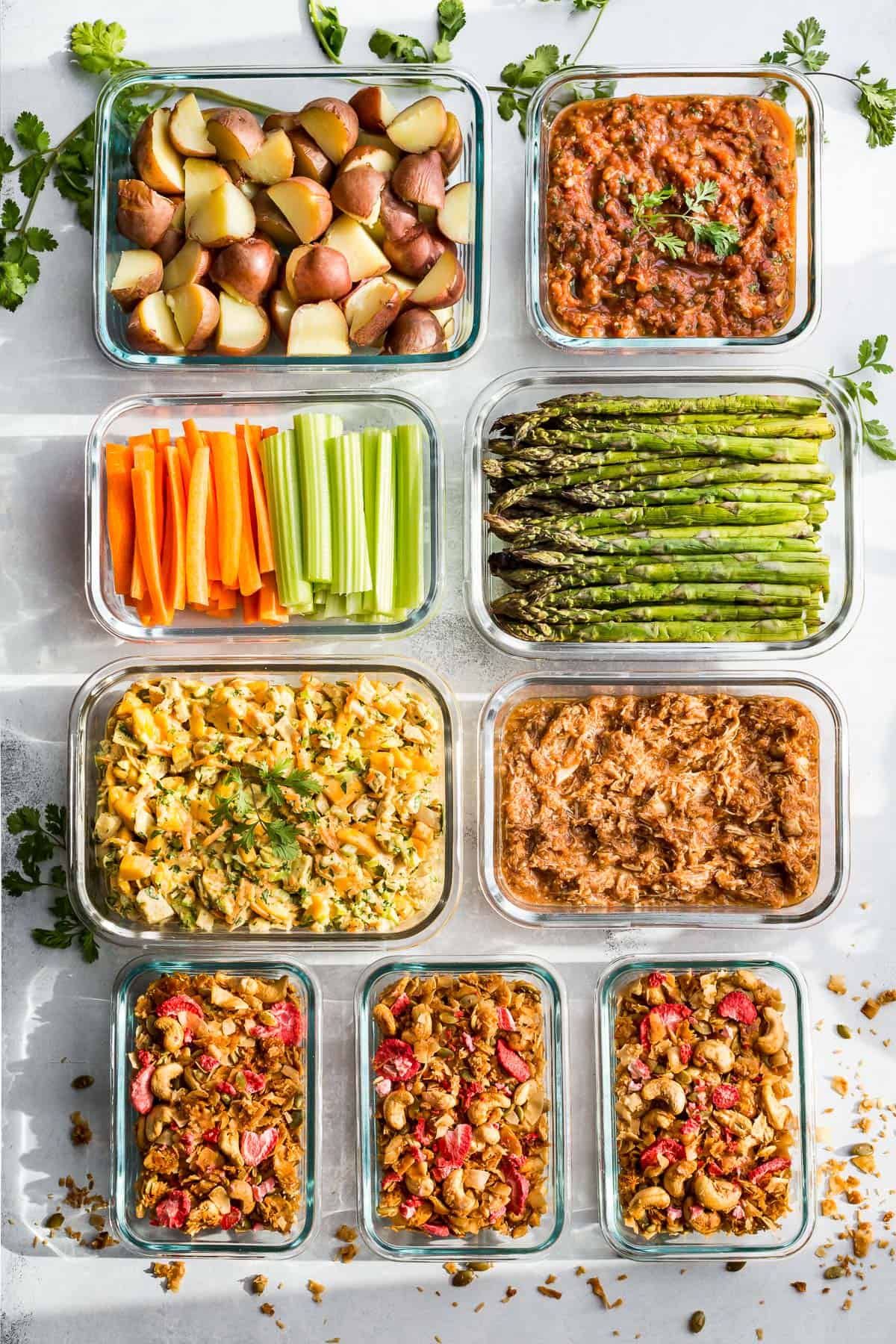 Glass meal prep containers filled with cooked potatoes, salsa, curried chicken salad, strawberry granola, chicken Tinga, and cooked asparagus.