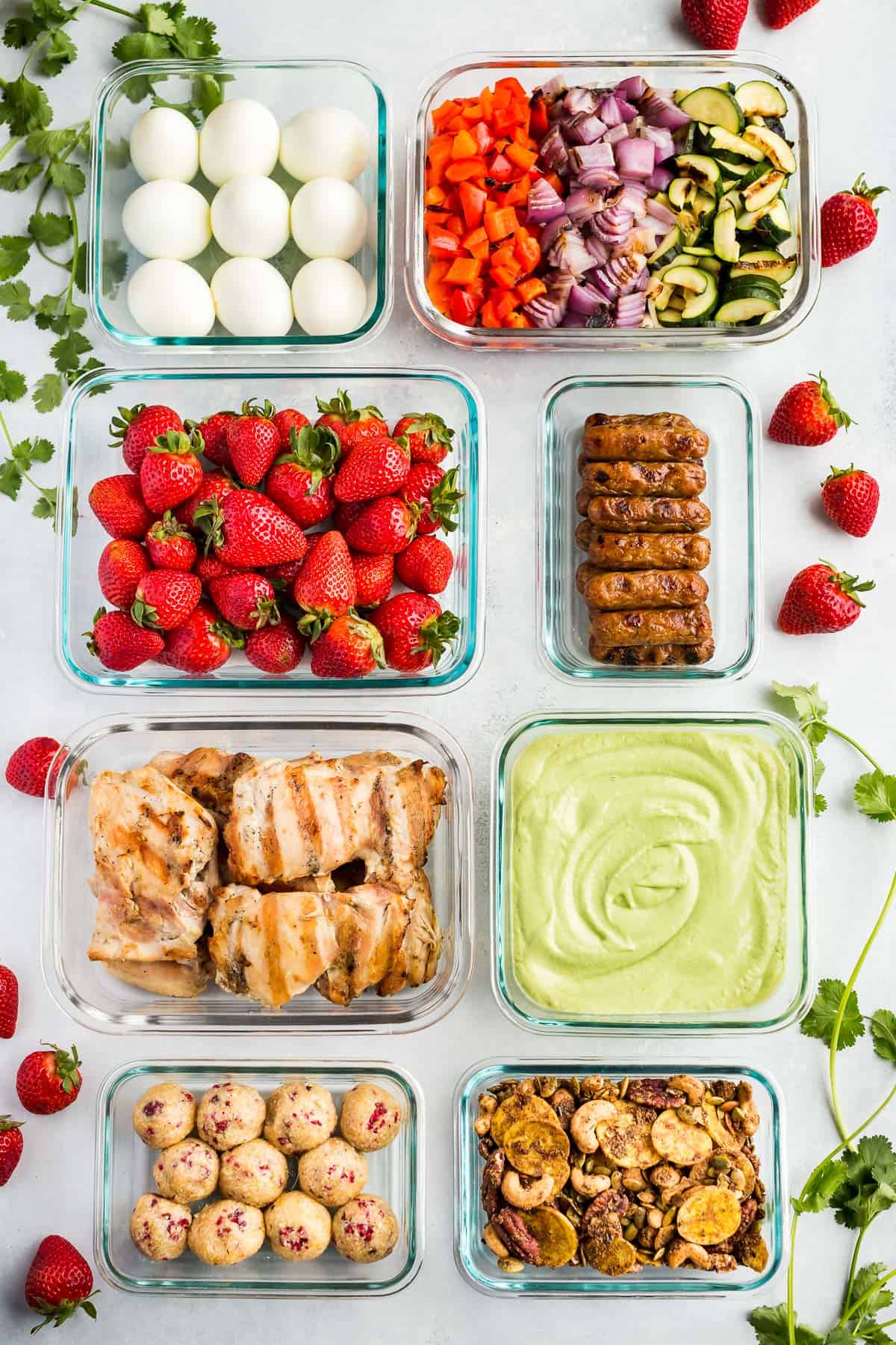 Glass meal prep containers filled with hardboiled eggs, grilled mixed veggies, strawberries, breakfast sausages, grilled chicken, energy bites, snack mix, and hummus.