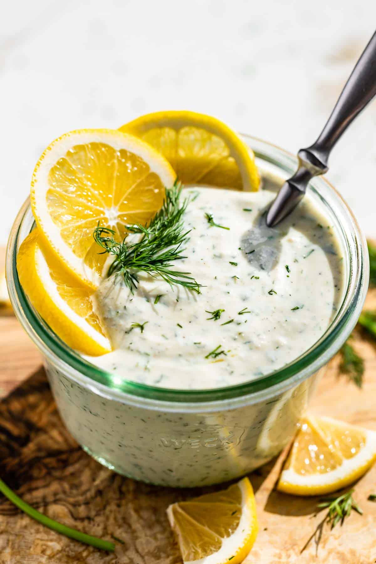 Greek yogurt dressing topped with a sprig of dill and lemon slices in a glass jar with a silver spoon in it.
