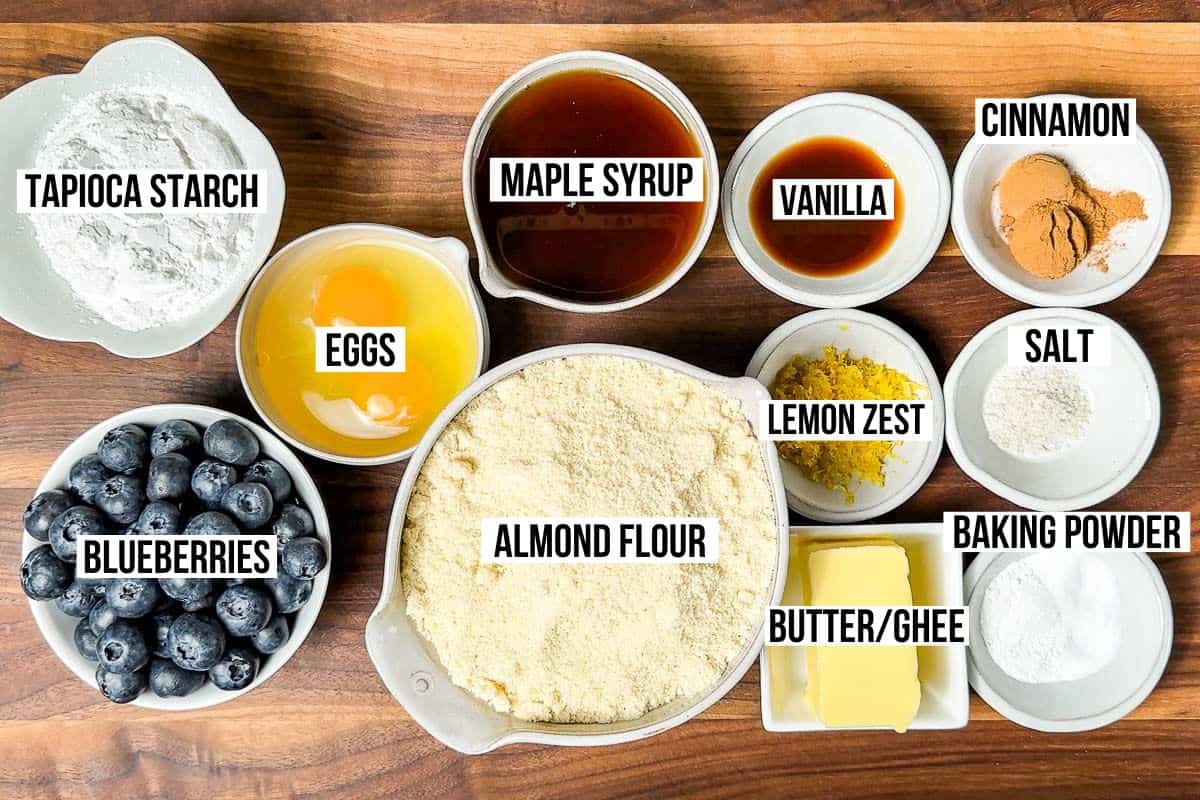 Almond flour, tapioca starch, eggs, maple syrup, vanilla extract, butter, baking powder, salt, cinnamon, and fresh blueberries in bowls on a wood cutting board.