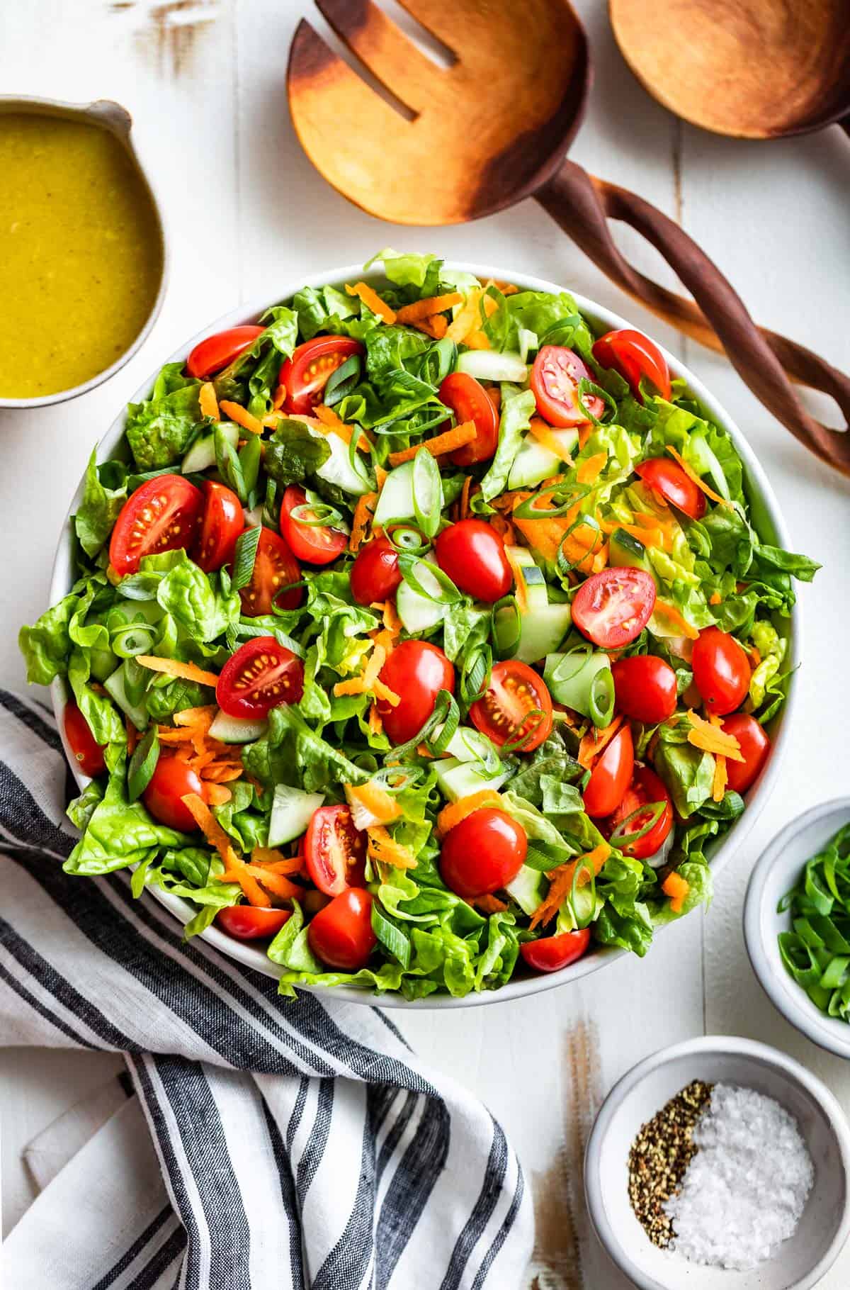Straight down view of Green Salad topped with tomatoes and cucumbers in a white bowl with wood spoons and vinaigrette on the side.