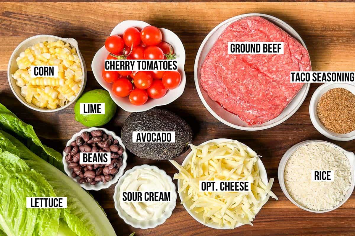 Lettuce, beans, ground beef, grated cheese, corn, cherry tomatoes, lime, avocado, taco seasoning, and rice in bowls on a wood cutting board.