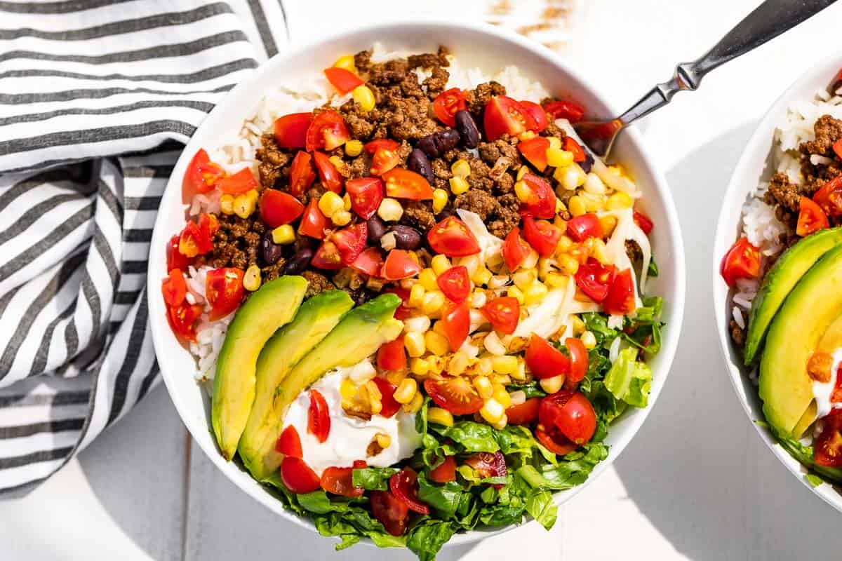 Straight down view of all the ingredients for taco bowls in two white bowls with a striped linen on the side.