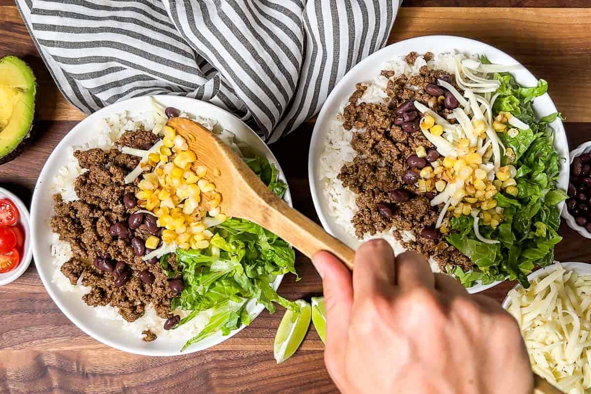 Spooning corn over the top of taco meat and lettuce in bowls of rice.