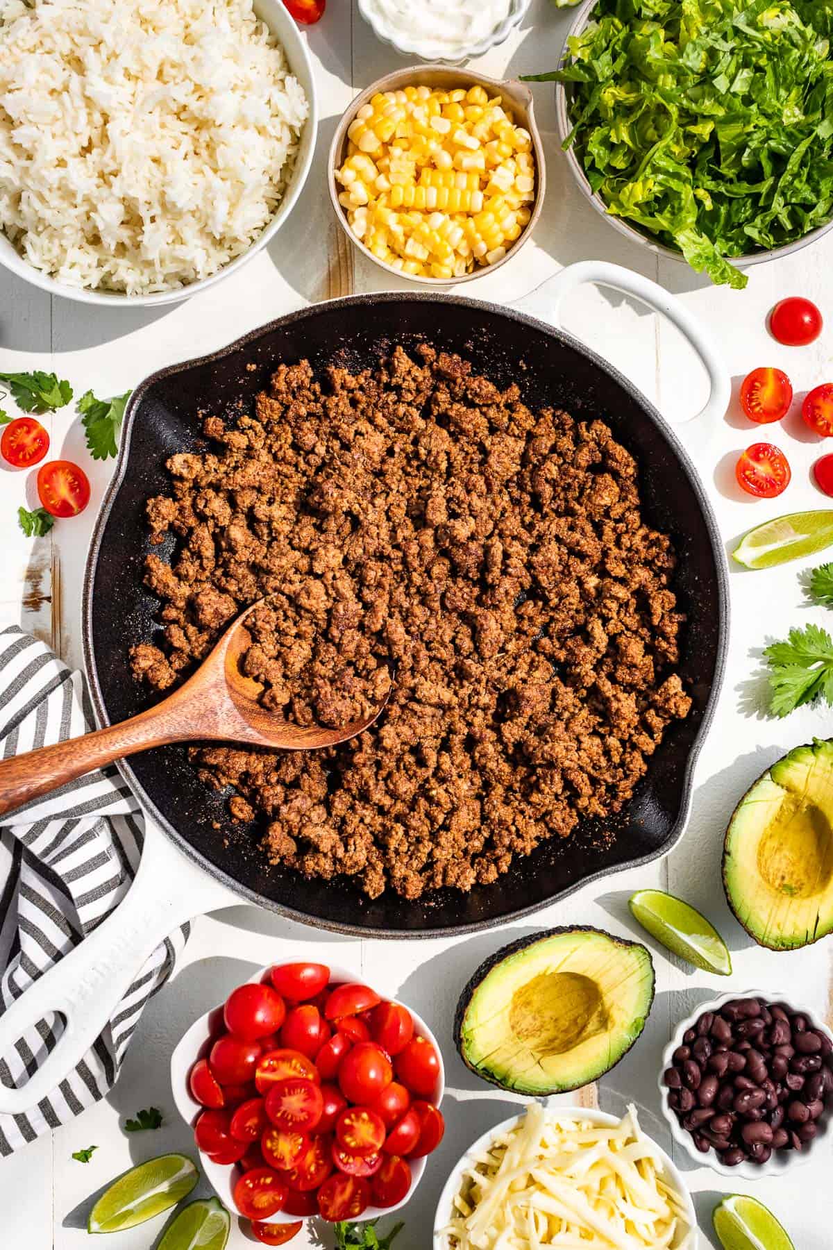 A large white skillet with taco meat surrounded by cherry tomatoes, avocados, grated cheese, shredded lettuce, beans and steamed rice in bowls.