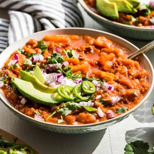 Side view of the finished Whole30 chili in two bowls with all the toppings and a halved avocado on the side.