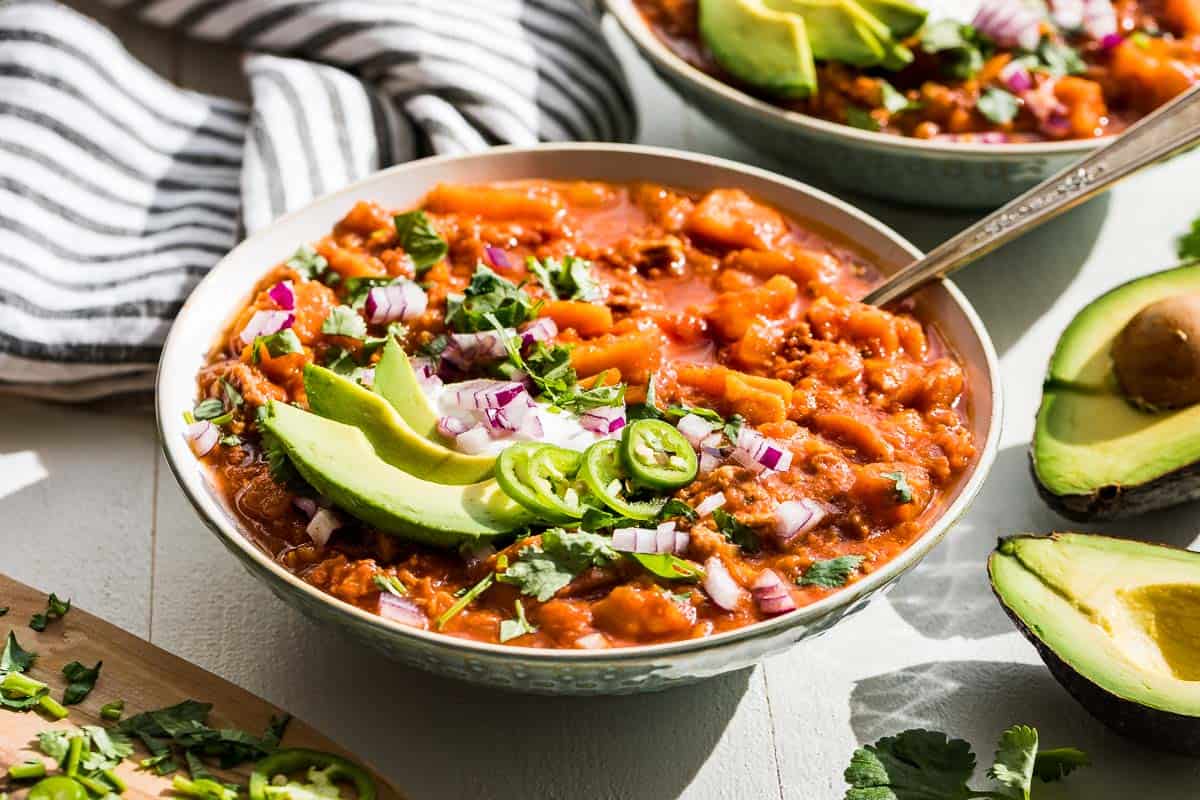 Side view of the finished Whole30 chili in two bowls with all the toppings and a halved avocado on the side.