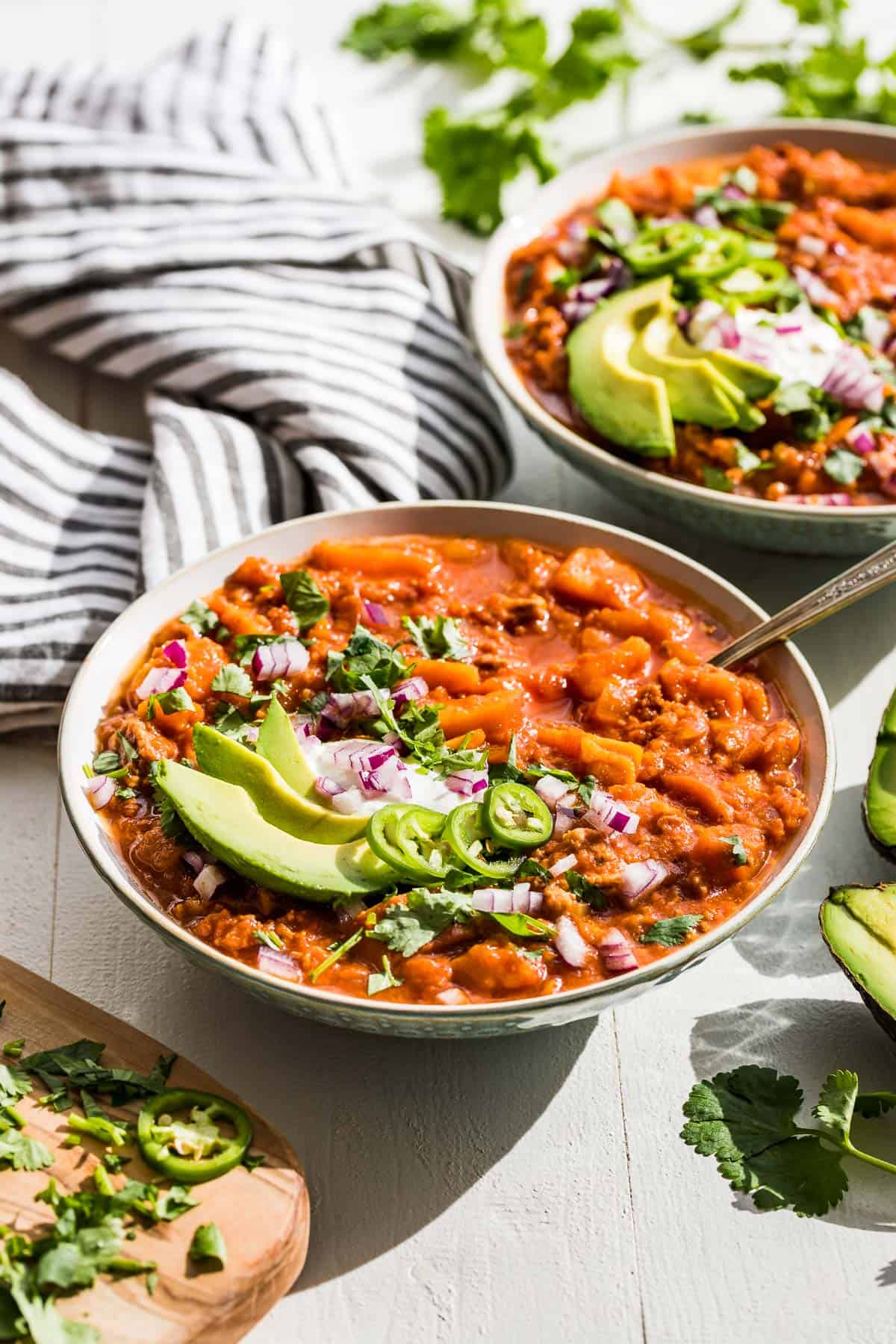 Two bowls of Whole30 chili with sliced avocado, jalapeno, red onion, and cilantro on top.