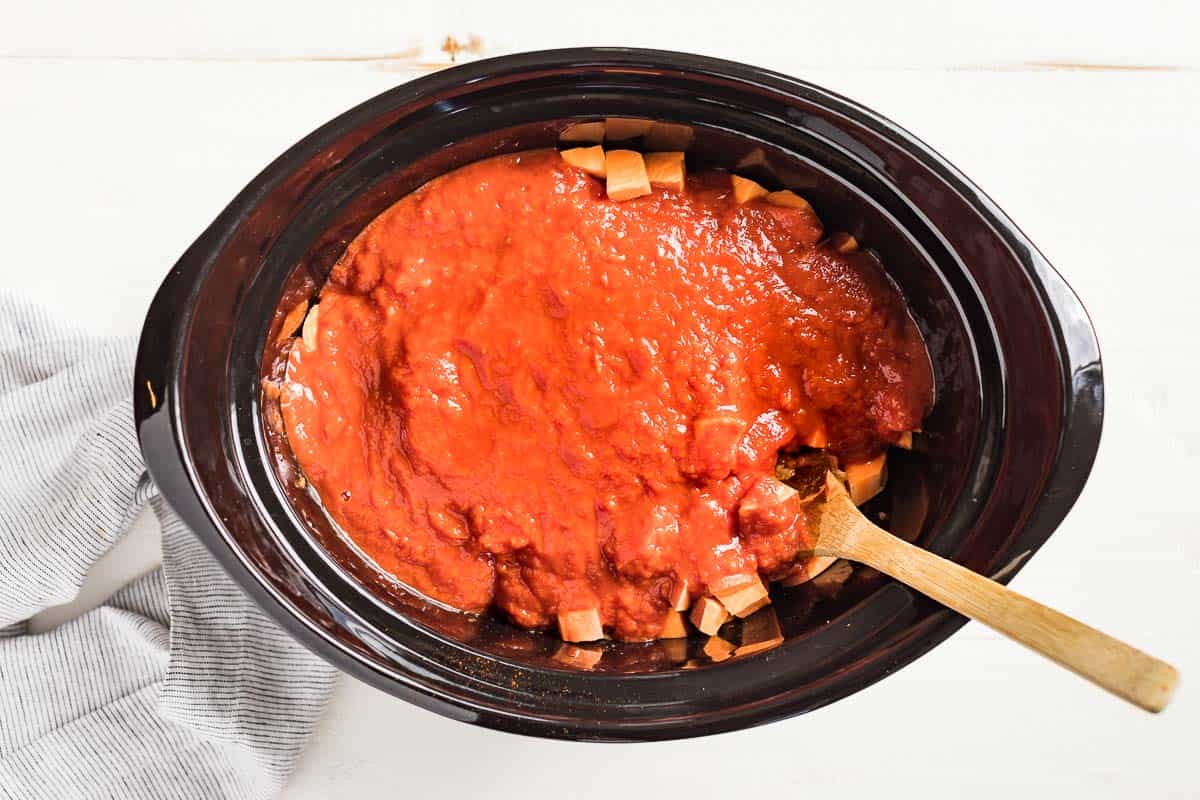 Pouring the tomato sauce over the top of the sweet potatoes in the slow cooker bowl.
