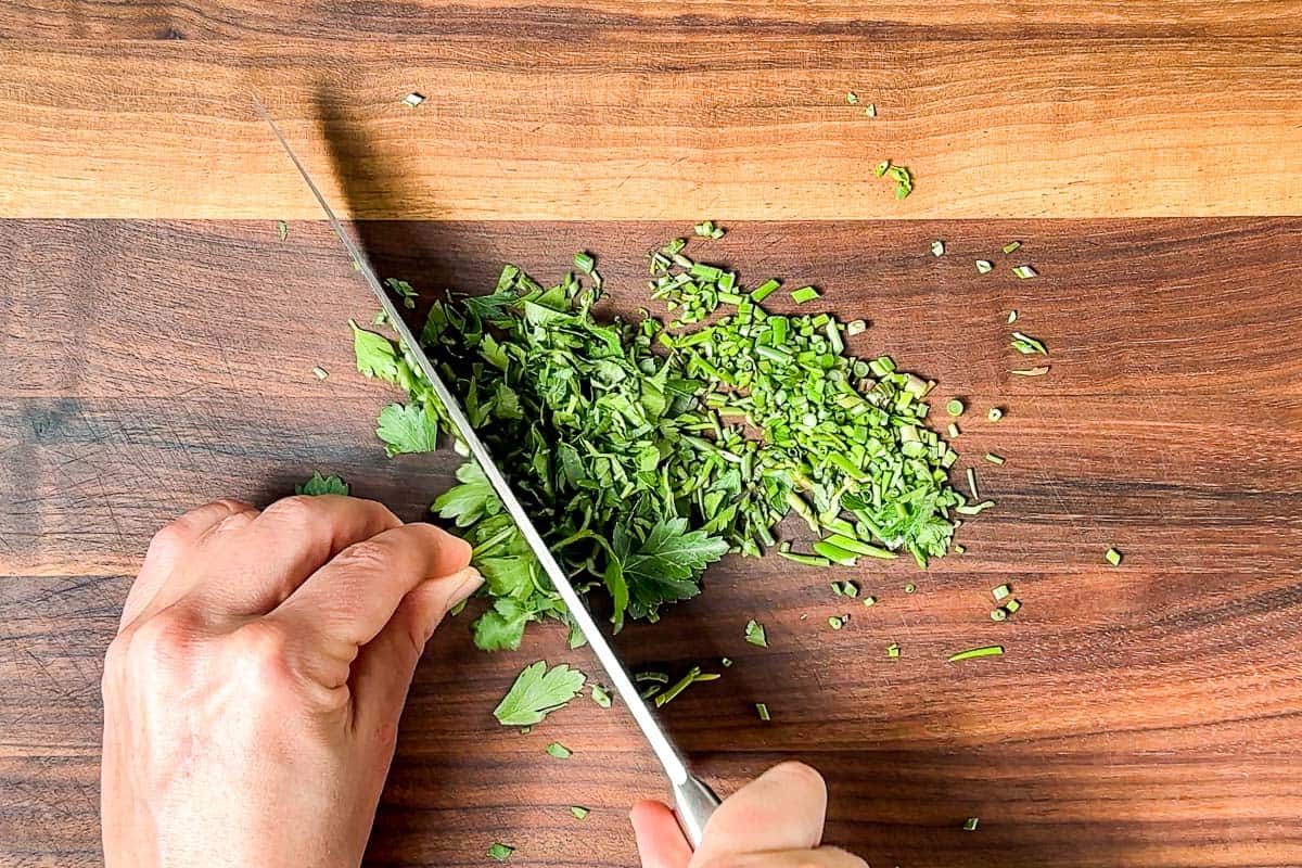 Mincing up chives and parsley with a chef's knife on a wood cutting board.
