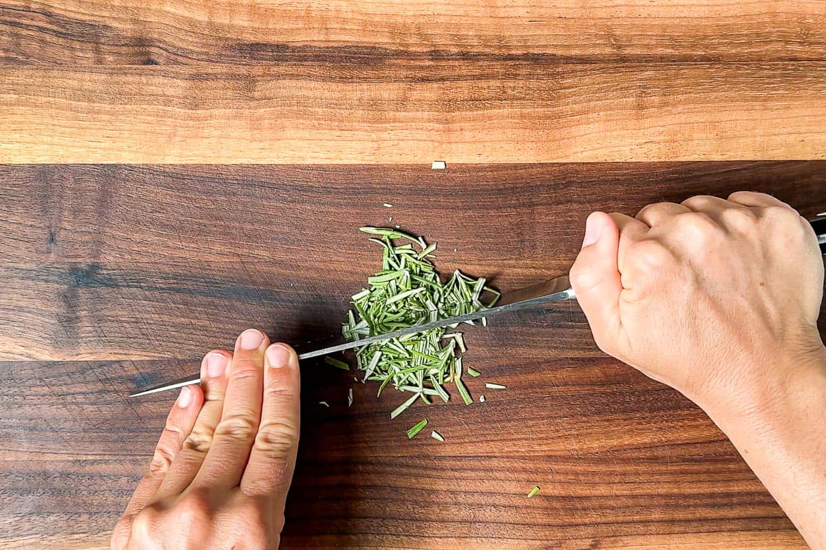 Two hands mincing up the fresh rosemary on a wood cutting board.