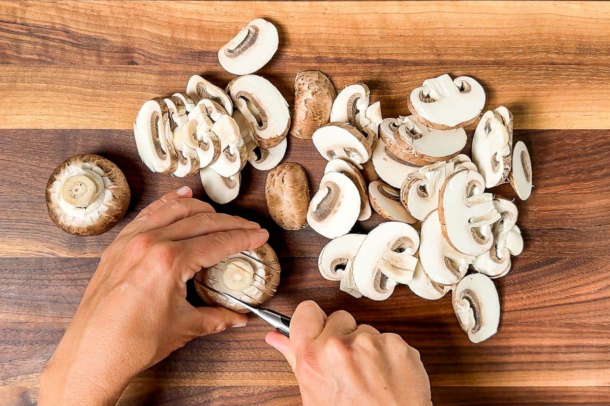Two hands using a pairing knife to slice up the Crimini mushrooms.