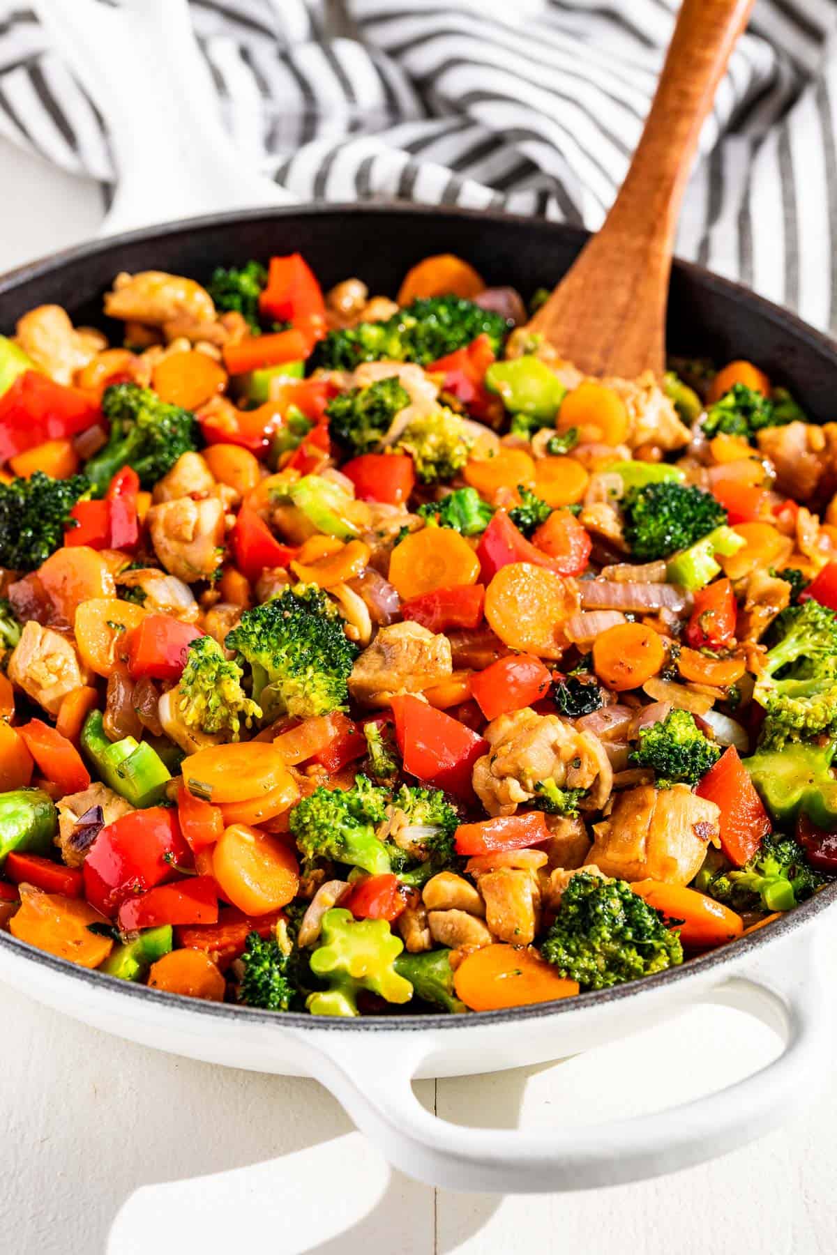 Honey Garlic Chicken Stir Fry and vegetables in a white skillet with a wood spoon in it on a white background.