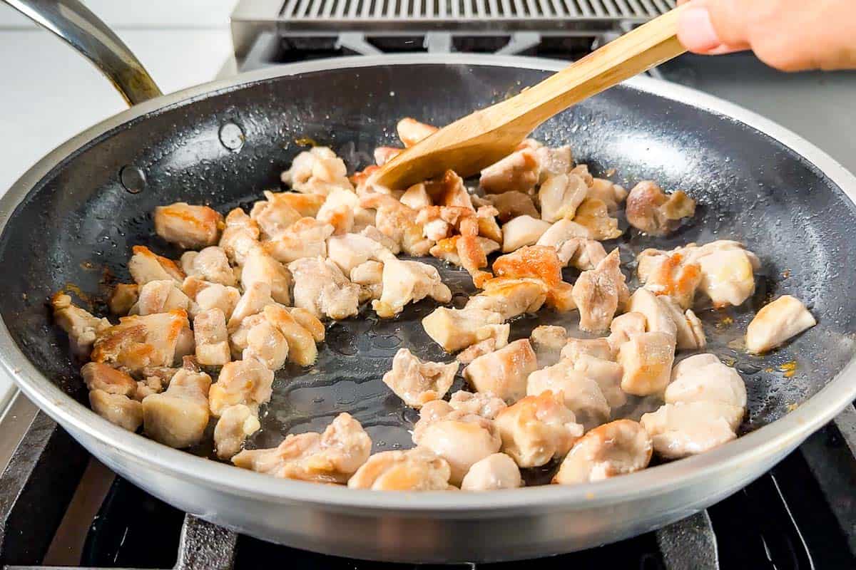 Sautéing the diced chicken thighs in a large skillet with a wood spoon on the stovetop.