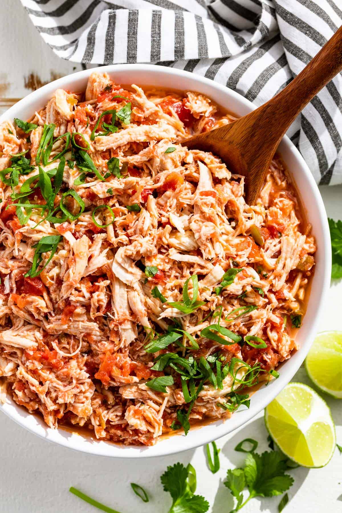 A large white bowl filled with shredded salsa chicken topped with sliced green onions and chopped cilantro with lime wedges on the side.