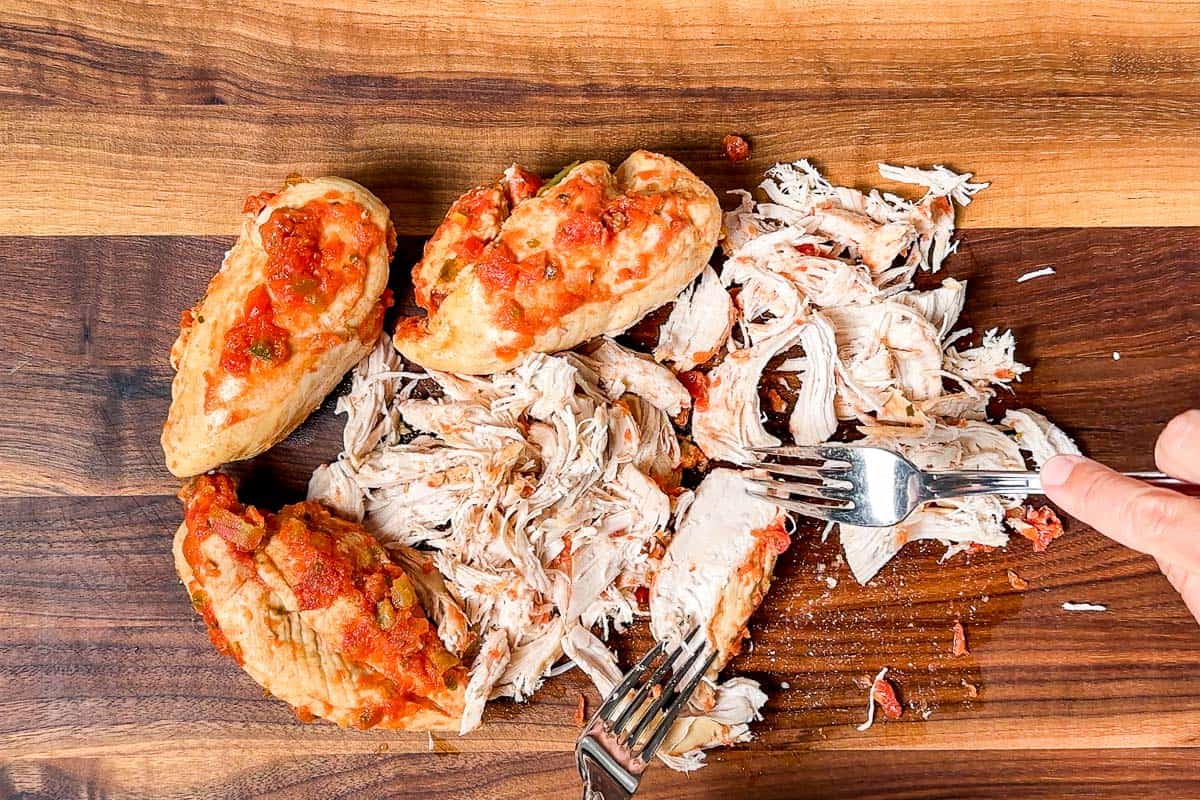 Shredding the cooked salsa chicken with two forks on a wood cutting board.