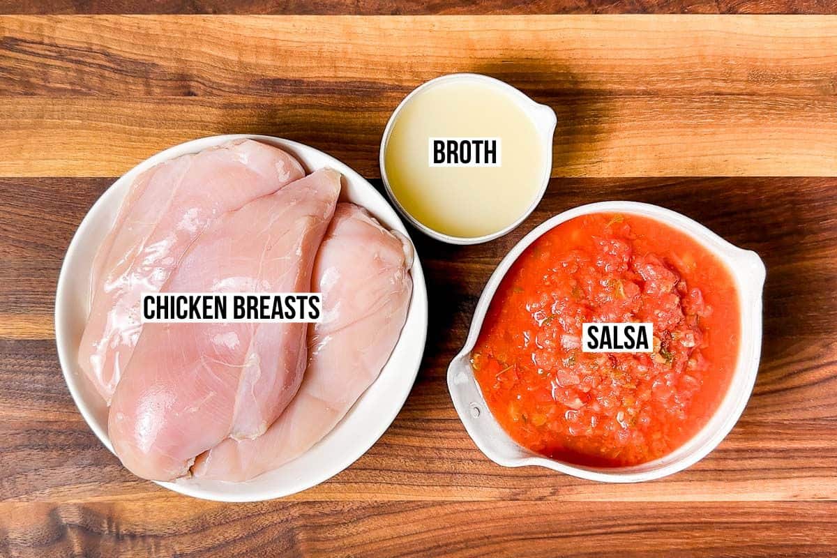 Chicken breasts, salsa, and chicken broth in bowls on a wood cutting board.