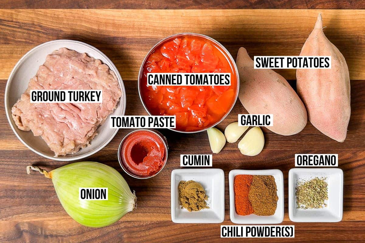 Ingredients for Whole30 chili: ground turkey, onion, garlic, chili powder, cumin, garlic, sweet potatoes, diced tomatoes, and tomato paste in small bowls on a wood cutting board.