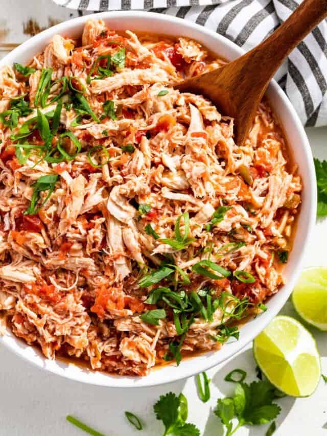 A large white bowl filled with shredded salsa chicken topped with sliced green onions and chopped cilantro with lime wedges on the side.