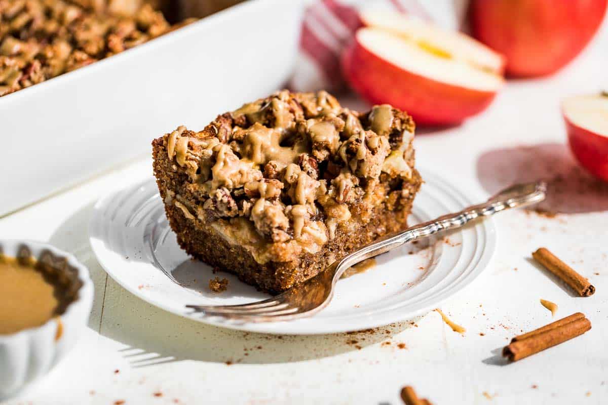 A large slice of Almond Flour Apple Cake on a white plate with a silver fork and apples on the side.